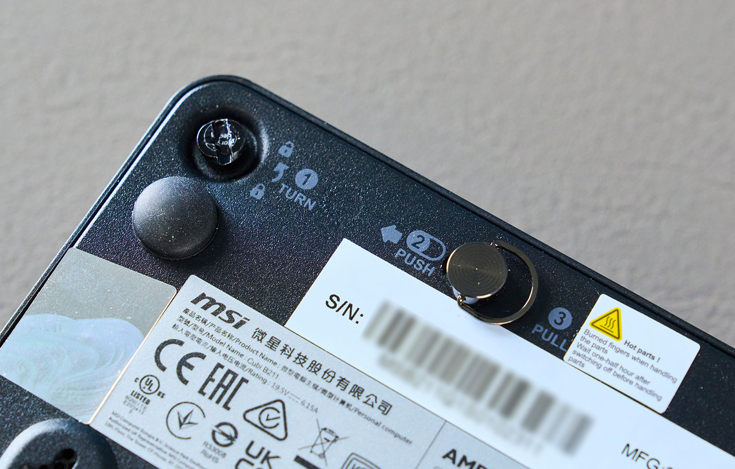 Cubi NUC AI+ 3MG 的機身底部也標示了拆卸底殼的方式，需要先將左上角的螺絲鬆開 (可用硬幣)，接著將卡榫向左推再向上拉開。